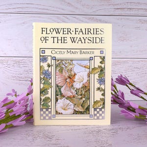 Könnte beinhalten: Ein gebundenes Buch mit dem Titel "Flower Fairies of the Wayside" von Cicely Mary Barker. Das Cover zeigt eine detaillierte Illustration einer Fee zwischen weißen Blumen und blauen Akzenten. Das Buch steht vor einem hellen Holzgrund mit lila Blumen.