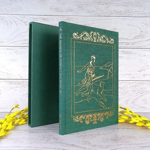 May include: A green hardcover book titled "The Masque of Comus" by John Milton. The cover features a gold illustration and ornate gold border. The book is displayed next to its matching green slipcase.