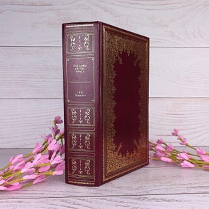 Puede incluir: Un libro de tapa dura color granate titulado The Lord of the Rings de J.R.R. Tolkien con detalles en pan de oro en el lomo y la portada. El lomo presenta volutas decorativas y nervios, mientras que la portada tiene un borde dorado ornamentado.