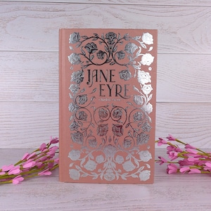 May include: A salmon-pink hardcover book titled "Jane Eyre". The cover is decorated with silver floral patterns and text. Pink flowers are arranged on either side of the book, on a wooden surface.