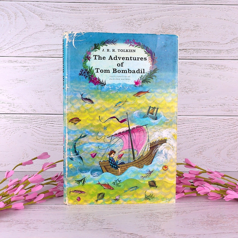 Op de afbeelding: Een vintage hardcover boek getiteld "The Adventures of Tom Bombadil" van J.R.R. Tolkien. De omslag toont een kleurrijke illustratie van een boot op een golvende zee, met vissen en een lezende persoon. De achtergrond is blauw en geel.