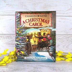 Pode incluir: Um livro de capa dura intitulado A Christmas Carol de Charles Dickens, ilustrado por Michael Foreman. A capa apresenta uma cena de inverno colorida e detalhada com uma árvore de Natal decorada, pessoas de cartola e uma rua nevada.