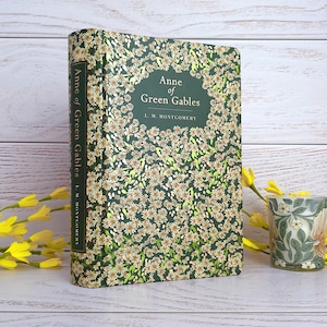 May include: A hardcover book titled "Anne of Green Gables" by L.M. Montgomery. The book's cover is dark green with a floral pattern and the title in a central frame. A small decorative cup with a floral design is next to the book.