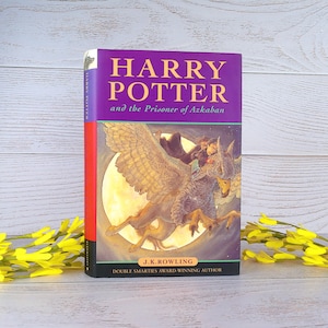 May include: A hardcover copy of "Harry Potter and the Prisoner of Azkaban" by J.K. Rowling. The book has a purple cover with gold lettering and a colorful illustration of a hippogriff. The book is a Double Smarties Award-Winning Author.