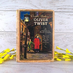 Pode incluir: Capa de livro vintage de "Oliver Twist" de Charles Dickens. A ilustração mostra duas figuras numa cena de rua, com o título e o nome do autor. O livro está envelhecido e apresenta sinais de desgaste.