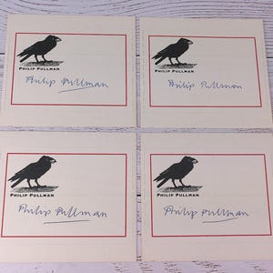 Peut inclure: Quatre cartes crème avec une illustration de corbeau noir et le nom "PHILIP PULLMAN" imprimé en noir. Chaque carte a une bordure rouge et une signature manuscrite à l'encre bleue "Philip Pullman".