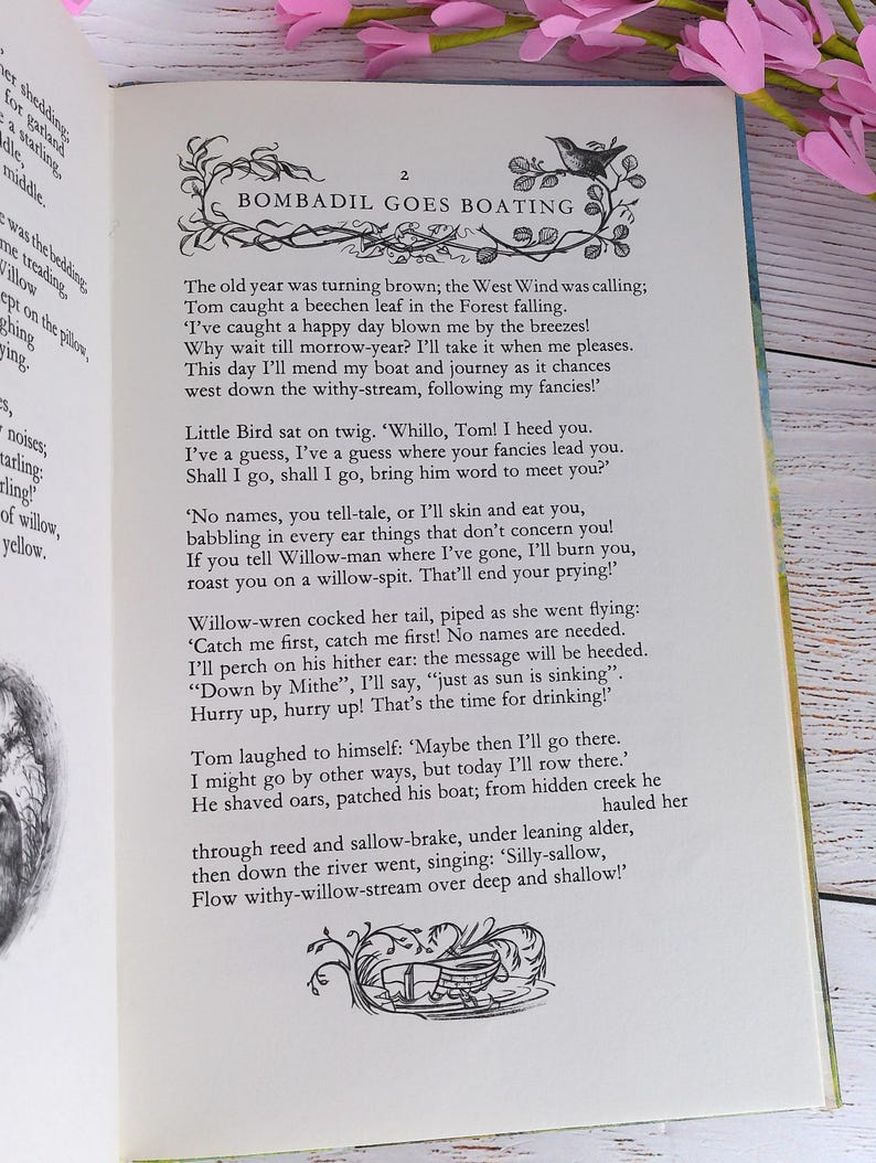 Op de afbeelding: Open boek met tekst en illustraties. De pagina toont de titel "Bombadil Goes Boating" en een decoratieve illustratie. De tekst beschrijft een verhaal over Tom en zijn reis. Roze bloemen zijn zichtbaar op de achtergrond.