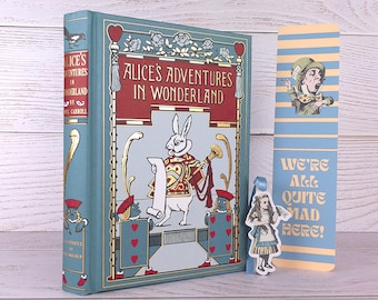 Puede incluir: Un libro de tapa dura titulado "Alicia en el país de las maravillas" con un marcapáginas a juego. El libro tiene una cubierta turquesa con detalles dorados e ilustraciones del Conejo Blanco y soldados de naipes. El marcapáginas presenta al Sombrerero Loco y el texto "We're All Quite Mad Here!"