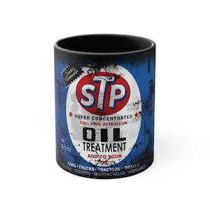 Könnte beinhalten: Eine schwarze Keramiktasse mit einer Vintage-Werbung für STP Ölbehandlung. Die Werbung zeigt einen roten Kreis mit dem STP-Logo, blauen und weißen Text und einen abgenutzten Hintergrund.