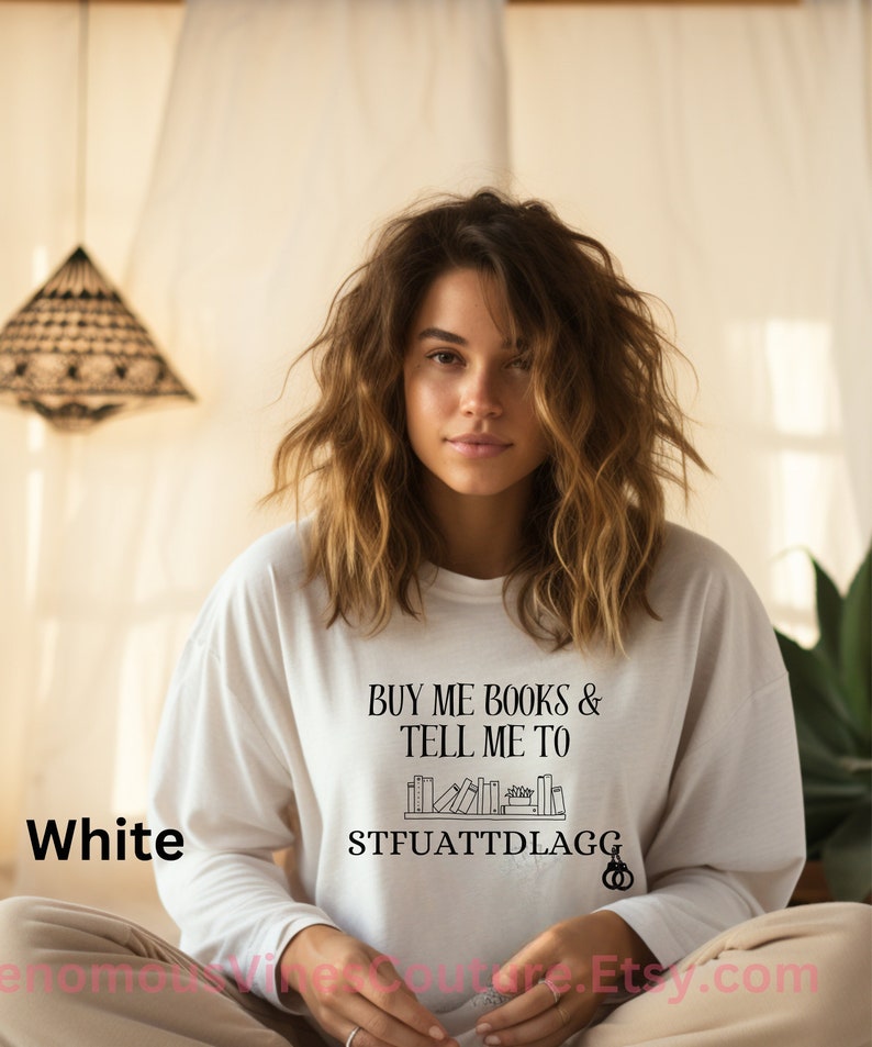 May include: White long-sleeve shirt with the text "Buy me books & tell me to STFUATTDLA GG" and a graphic of a bookshelf with books.