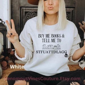 May include: White sweatshirt with the text "BUY ME BOOKS & TELL ME TO STFUATTDLAGG" and a graphic of a bookshelf with books on it.