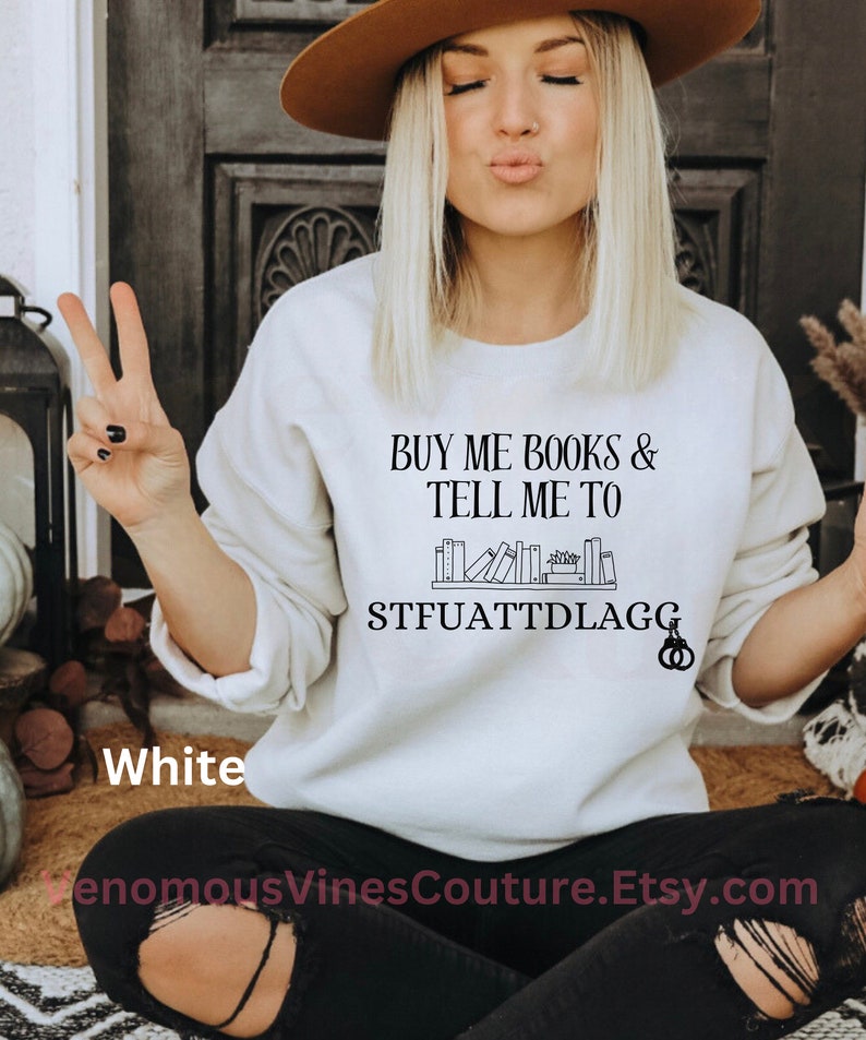 May include: White sweatshirt with the text "BUY ME BOOKS & TELL ME TO STFUATTDLAGG" and a graphic of a bookshelf with books on it.