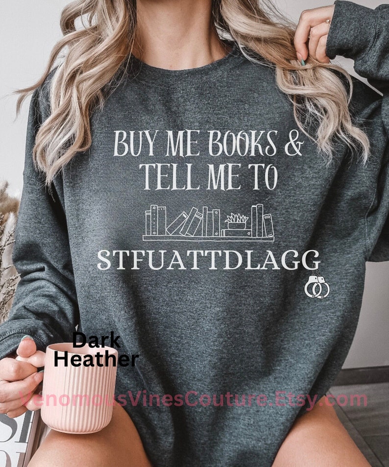 May include: Dark heather grey sweatshirt with the text "Buy me books & tell me to STFUATTDLAGG" in white. The sweatshirt features a graphic of a bookshelf with books and a plant.