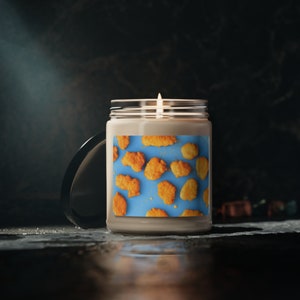 May include: A glass candle jar with a blue and white label featuring a pattern of fried chicken nuggets. The candle is lit and the jar has a black lid.