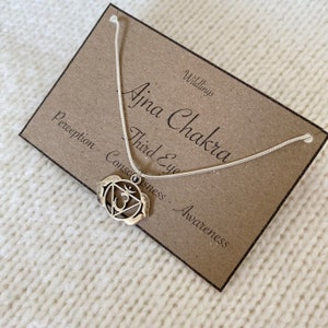 Op de afbeelding: Een zilverkleurige ketting met een hanger met het Ajna Chakra-symbool. Het sieraad wordt gepresenteerd op een rechthoekige bruine kaart met de tekst Wildlings, Ajna Chakra, Third Eye, Perception, Consciousness en Awareness erop gedrukt.