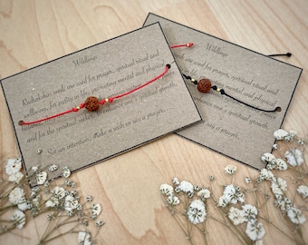 May include: Two Wildlings bracelets with Rudraksha seed beads on red and black cords. Each bracelet is displayed on a brown card with text about prayer and spiritual growth. White flowers are scattered around the cards.
