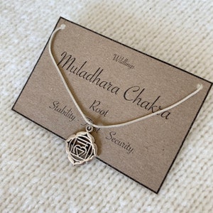 May include: A silver-toned necklace featuring a Muladhara Chakra pendant on a thin chain. The pendant is displayed on a rectangular brown card printed with the text Muladhara Chakra, Wildlings, Stability, Root, and Trust - Security.