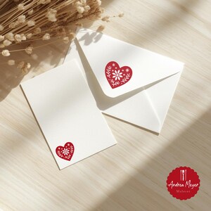 May include: White stationery set featuring a red heart design with floral accents. The set includes a blank card and an envelope, both adorned with the same heart motif. The design is a folk art style. The stationery is on a light wood surface.