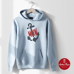 May include: A light blue hoodie with a front pocket and a drawstring hood. The hoodie features a graphic of a dark gray anchor with the word "MOIN" in red letters. The hoodie is hanging on a wooden hanger.