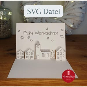 Peut inclure: Carte de Noël blanche en pop-up avec une scène de village, comprenant des maisons, une église et un arbre. La carte porte les mots "Frohe Weihnachten" et des illustrations d'étoiles. Le texte "SVG Datei" est en haut.