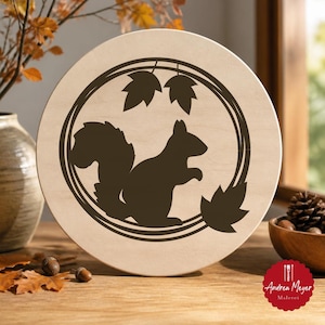 May include: A circular light-colored wooden plaque featuring a dark brown laser-cut silhouette of a squirrel centered within a circular frame. The design includes three stylized maple leaves positioned at the top and bottom of the circular border.