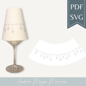 Op de afbeelding: Een witte lampenkap met een hartjesontwerp, rustend op een wijnglas. Het ontwerp heeft kleine, hangende hartjes. Er wordt ook een apart hartjesslingerontwerp getoond. De tekst "PDF SVG" is inbegrepen.