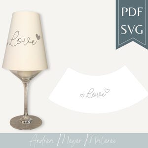 Può includere: Un paralume color crema con la parola "Love" in corsivo e un cuore. Il paralume è su uno stelo e una base di vetro trasparente. Viene mostrato anche un ritaglio di carta bianca separato con la parola "Love" e un cuore.