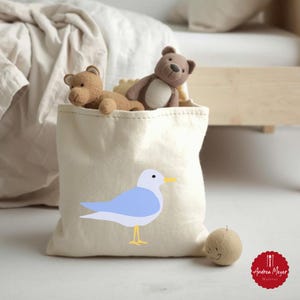 以下が含まれることがあります： 青いカモメのグラフィックが入ったベージュのキャンバス収納バッグ。茶色のテディベアが2つ入っています。小さな丸いベージュのフェルトボールには笑顔が描かれています。バッグはベッドの前にあります。