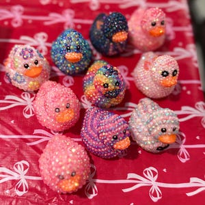 May include: A collection of bedazzled duck figurines in various colors, including pink, blue, and rainbow hues. Each duck has an orange beak and is covered in sparkling embellishments. The ducks are arranged on a red surface.
