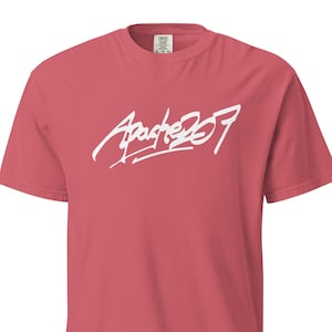 Könnte beinhalten: Korallfarbenes T-Shirt mit kurzen Ärmeln und dem weißen Schriftzug "Apache 207" in einem stilisierten Schriftzug auf der Brust. Das T-Shirt hat einen Rundhalsausschnitt und eine lockere Passform.