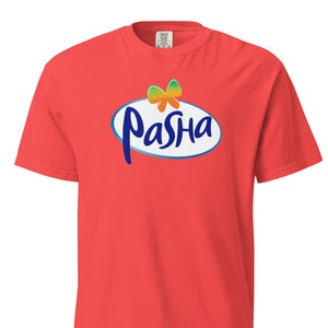 Könnte beinhalten: Ein korallenfarbenes T-Shirt mit dem Wort "PaSha" in blauer Schrift in einem weißen Oval mit blauem Rand. Eine farbenfrohe Bogen-Grafik befindet sich über dem Text. Das Shirt besteht aus einem weichen Material.