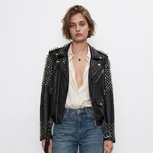 May include: A black leather motorcycle jacket featuring silver-toned metal studs on the shoulders and sleeves, paired with a light-coloured button-down shirt, blue denim jeans, and black loafers. The jacket includes a notched collar and a belt.
