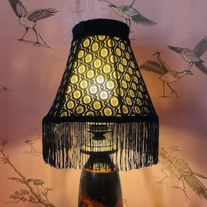 May include: A table lamp featuring a dark, amber-toned glass base and a black lampshade with a geometric, lace-like pattern and long, dangling black fringe. The lamp emits a warm, soft glow from within the shade, illuminating the intricate design.