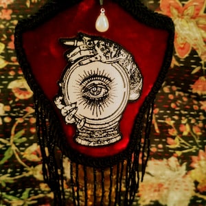Puede incluir: Un adorno de pared en forma de escudo de terciopelo rojo con ribete y flecos negros. Presenta un parche con una mano sosteniendo una bola de cristal con un diseño de ojo. Una perla nacarada cuelga de la parte superior.