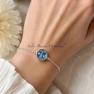 May include: A silver-toned chain bracelet featuring a circular pendant filled with small, textured blue and white fragments. The text Salty Beach Memorial is visible above the pendant. The bracelet has a delicate link design and a metallic finish.