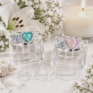 May include: Two silver-toned rings featuring heart-shaped centerpieces with iridescent flecks. One ring has a teal heart, the other a pale pink heart. Both rings have small wing-shaped accents on the sides and are displayed on clear stands.