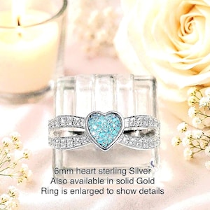 May include: A sterling silver ring featuring a 6mm heart-shaped centerpiece filled with blue glittery material, flanked by bands encrusted with small clear stones. Text on the image reads: 6mm heart sterling Silver. Also available in solid Gold. Ring is enlarged to show details.