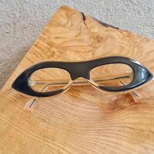 May include: Black cat-eye glasses with gold metal arms on a wooden surface.
