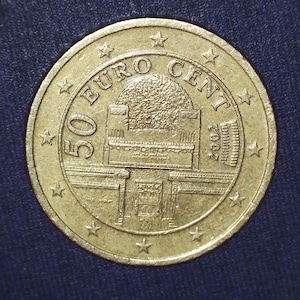 May include: A gold-colored 50 Euro Cent coin from 2002. The coin features an architectural design in the center, surrounded by the words "50 EURO CENT" and star-shaped symbols. The coin is set against a dark blue background.