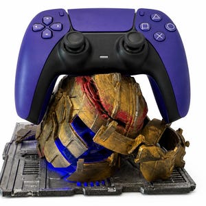 Puede incluir: Un mando de videojuegos morado y negro descansa sobre un casco dorado desgastado con detalles rojos. El casco se asienta sobre una base rectangular de aspecto metálico con un patrón de rejilla y una luz azul brillante debajo.