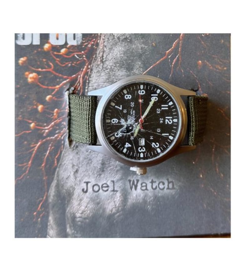K&ouml;nnte beinhalten: Eine silberne Armbanduhr mit schwarzem Zifferblatt, wei&szlig;en Ziffern und Zeigern sowie einem roten Sekundenzeiger. Sie hat ein gr&uuml;nes Stoffarmband. Der Text "Joel Watch" ist darunter zu sehen.