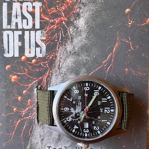 K&ouml;nnte beinhalten: Eine silberne Armbanduhr mit schwarzem Zifferblatt und gr&uuml;nem Canvas-Armband. Das Zifferblatt zeigt wei&szlig;e Ziffern und Zeiger, mit dem Text "Joel Watch" unten. Das Zifferblatt ist gesprungen. Der Hintergrund zeigt den Text "THE LAST OF US".