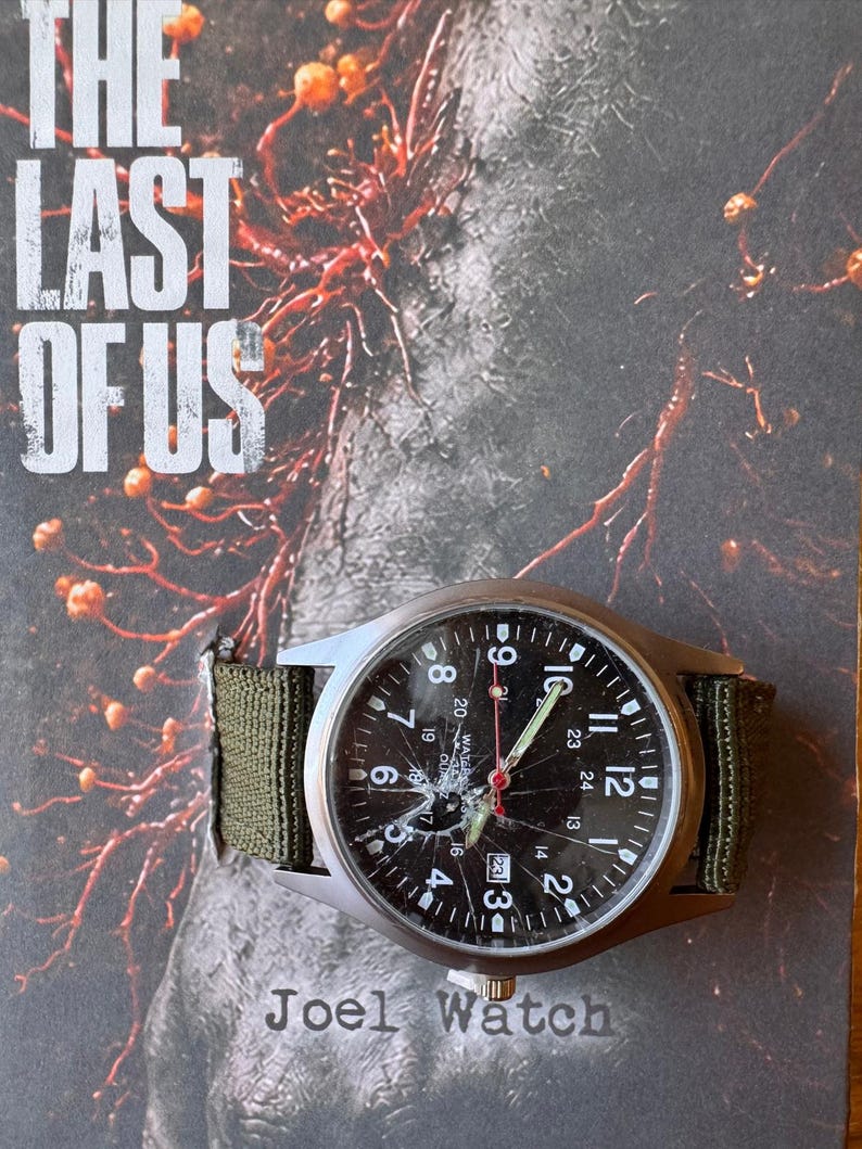 K&ouml;nnte beinhalten: Eine silberne Armbanduhr mit schwarzem Zifferblatt und gr&uuml;nem Canvas-Armband. Das Zifferblatt zeigt wei&szlig;e Ziffern und Zeiger, mit dem Text "Joel Watch" unten. Das Zifferblatt ist gesprungen. Der Hintergrund zeigt den Text "THE LAST OF US".