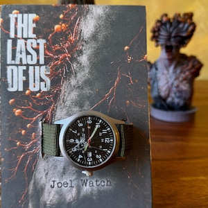 K&ouml;nnte beinhalten: Eine Armbanduhr mit schwarzem Zifferblatt und wei&szlig;en Ziffern, mit einem gr&uuml;nen Canvas-Armband. Die Uhr wird auf einem Buchcover mit dem Text "THE LAST OF US" und "Joel Watch" pr&auml;sentiert. Eine Figur ist im Hintergrund zu sehen.