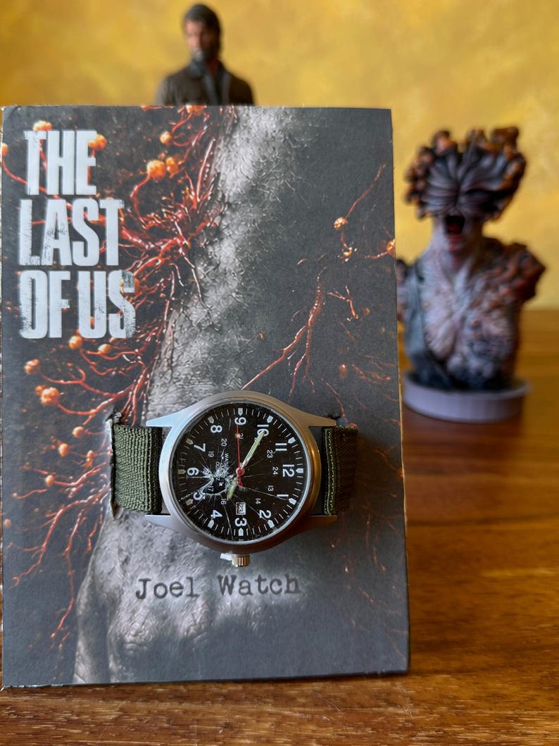 K&ouml;nnte beinhalten: Eine Armbanduhr mit schwarzem Zifferblatt und wei&szlig;en Ziffern, mit einem gr&uuml;nen Canvas-Armband. Die Uhr wird auf einem Buchcover mit dem Text "THE LAST OF US" und "Joel Watch" pr&auml;sentiert. Eine Figur ist im Hintergrund zu sehen.