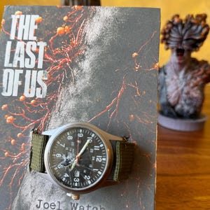 K&ouml;nnte beinhalten: Eine Armbanduhr mit schwarzem Zifferblatt und gr&uuml;nem Canvas-Armband wird auf einem Buchcover pr&auml;sentiert. Die Uhr hat ein silbernes Geh&auml;use und sichtbare Zahlen. Das Buchcover tr&auml;gt den Titel "THE LAST OF US" und den Text "Joel Watch".