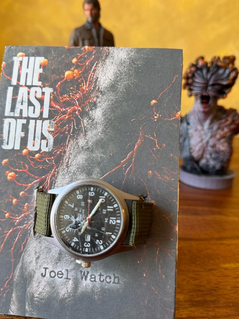 K&ouml;nnte beinhalten: Eine Armbanduhr mit schwarzem Zifferblatt und gr&uuml;nem Canvas-Armband wird auf einem Buchcover pr&auml;sentiert. Die Uhr hat ein silbernes Geh&auml;use und sichtbare Zahlen. Das Buchcover tr&auml;gt den Titel "THE LAST OF US" und den Text "Joel Watch".