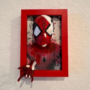 Puede incluir: Obra de arte enmarcada que presenta una máscara de Spider-Man roja y blanca con detalles negros en los ojos. La obra está enmarcada en un marco rojo, con una mano roja a juego que se extiende desde el marco. La obra tiene una apariencia texturizada.