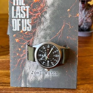 K&ouml;nnte beinhalten: Eine silberfarbene Armbanduhr mit schwarzem Zifferblatt und wei&szlig;en Ziffern, an einem gr&uuml;nen Canvas-Armband. Die Uhr wird auf einer Karte mit dem Text "THE LAST OF US" und "Joel Watch" pr&auml;sentiert.