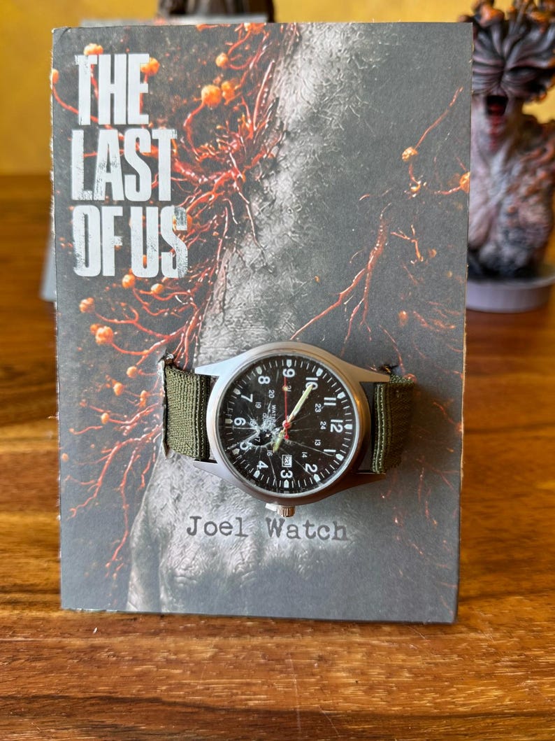 K&ouml;nnte beinhalten: Eine silberfarbene Armbanduhr mit schwarzem Zifferblatt und wei&szlig;en Ziffern, an einem gr&uuml;nen Canvas-Armband. Die Uhr wird auf einer Karte mit dem Text "THE LAST OF US" und "Joel Watch" pr&auml;sentiert.